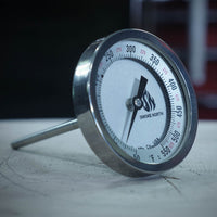 A Tel Tru offset smoker thermometer with custom 'Smoke North' logo shown on top of a wood splitting log