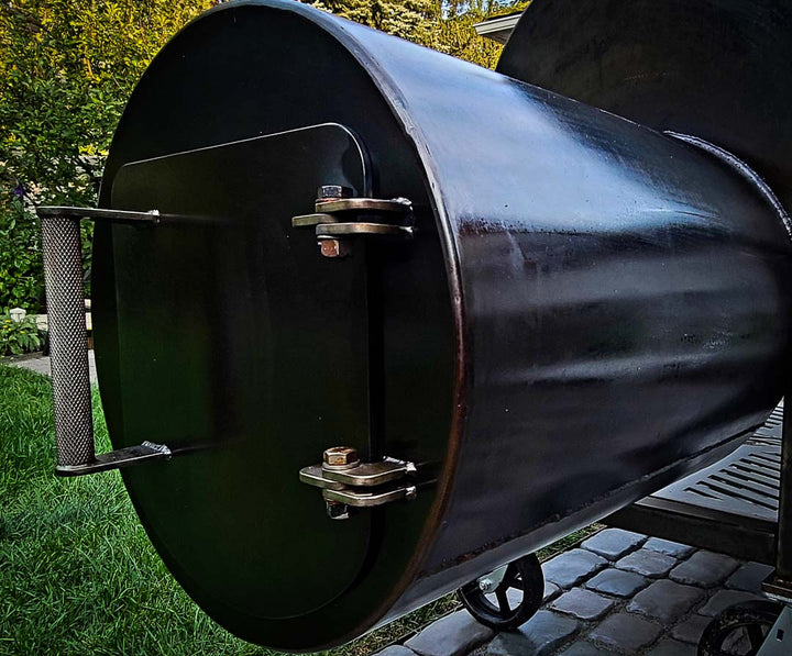 An offset smoker firebox with a dark oiled finish, a knurled stainless steel door handle, and stainless steel hinges shown outdoors on a patio